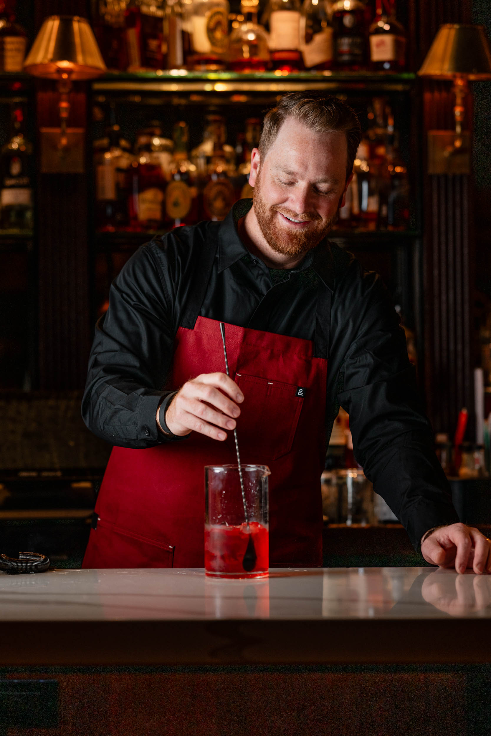 Bartender stirring a cocktail behind the bar in a vibrant red apron