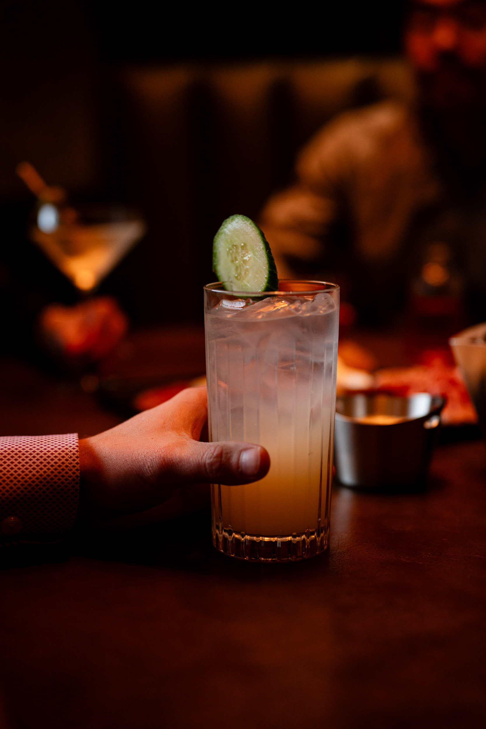 man hand extended onto the table holding onto his cocktail in a tall glass with a cucumber garnish